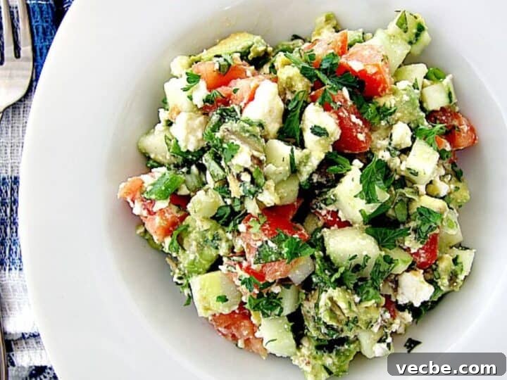 A bowl of avocado salad containing tomatoes, cucumbers, feta cheese, and herbs, presented on a white background.