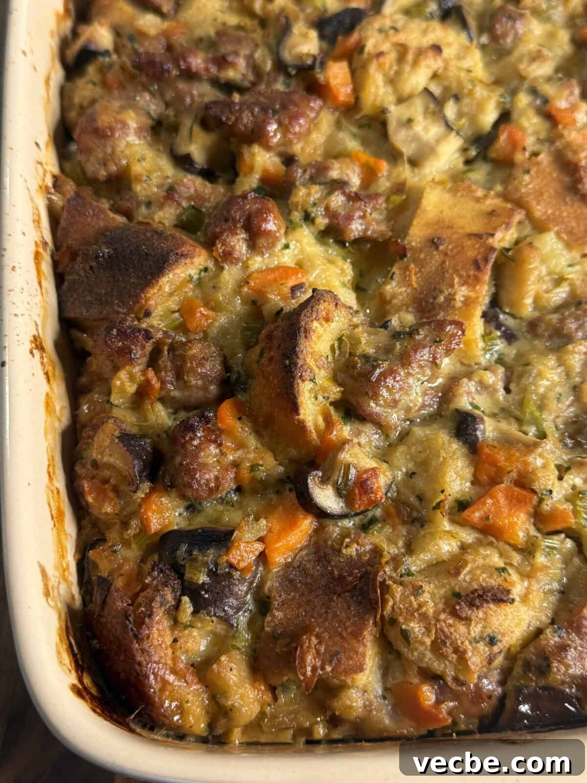 Sausage and Leek Bread Pudding-Style Stuffing 3 A close-up of a baked casserole in a rectangular dish, featuring chunks of bread, sausage, carrots, mushrooms, and herbs, with a golden-brown, crispy top.