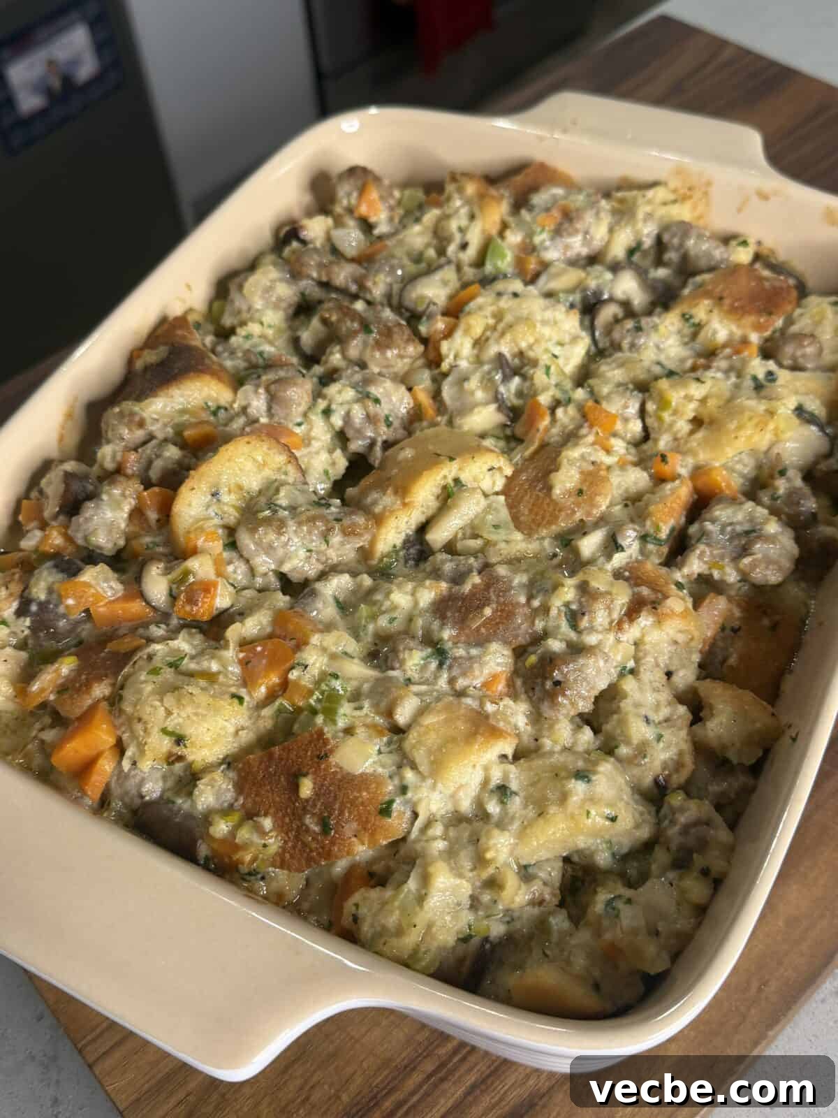 Sausage and Leek Bread Pudding-Style Stuffing 4 A casserole dish filled with homemade stuffing, featuring chunks of bread, sausage, carrots, mushrooms, and herbs, sitting on a wooden surface in a sunlit kitchen.