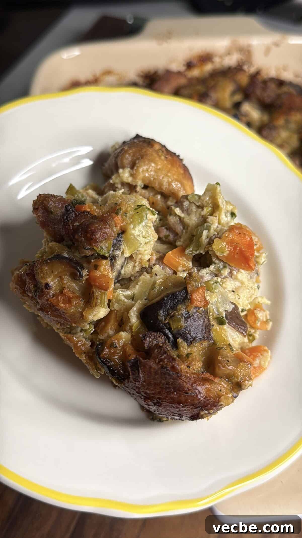 Sausage and Leek Bread Pudding-Style Stuffing 5 A close-up of a serving of vegetable bread pudding, featuring chunks of bread, carrots, celery, eggplant, and other mixed vegetables on a white plate with a yellow rim.