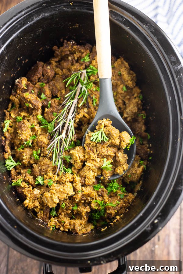 Herbed Slow Cooker Stuffing for Holiday Dinners 3 Large oval slow cooker crock filled with crockpot stuffing topped with fresh parsley and a sprig of rosemary with a wooden and silicone spoon in the middle on a rustic wood background.